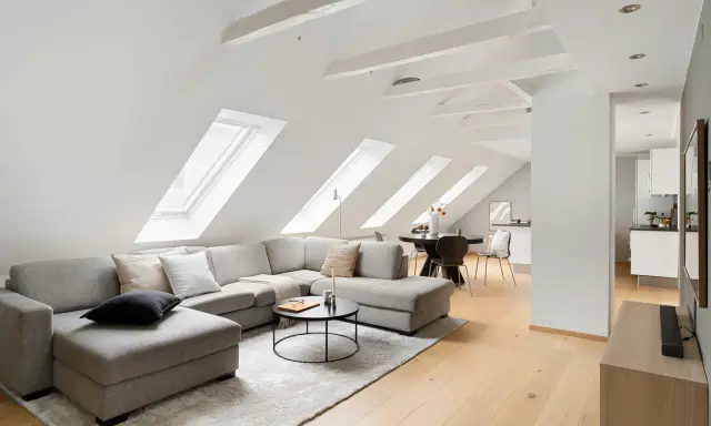 Soggiorno mansardato con divano angolare grigio, tavolino nero e travi in legno a vista sul soffitto inclinato.