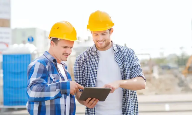 Due operai con caschi gialli esaminano un tablet, forse per un verbale di sopralluogo cantiere. Sullo sfondo, un cantiere con bagni chimici.
