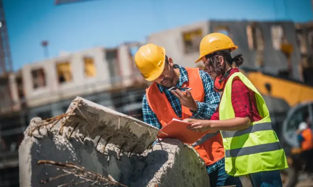 Due operai con caschi gialli e giubbotti ad alta visibilità discutono un progetto di **construction management** su un cantiere.