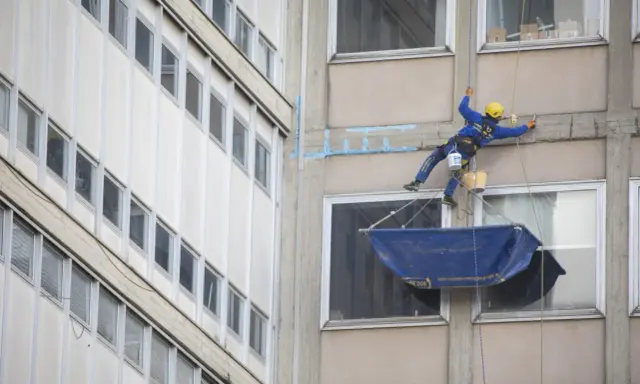 Operaio in corda esegue consolidamenti strutturali su un edificio, con attrezzature di sicurezza e secchi.