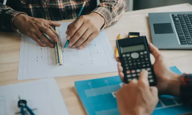 Due mani lavorano su progetti, una usa una calcolatrice, l'altra una matita e un righello, per definire i requisiti igienico sanitari.