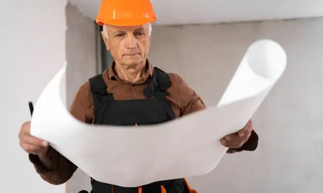 Un esperto capomastro, con casco arancione, studia attentamente un progetto, dimostrando il vero significato di guida e competenza in cantiere.