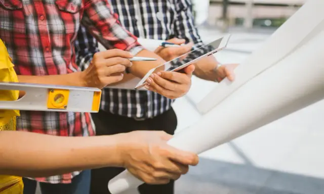 Team di operai esamina progetto su tablet, livella e rotoli di carta. Aggiornamento sal stato avanzamento lavori.