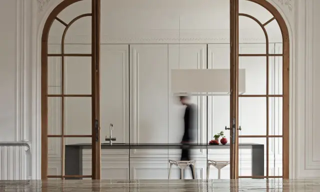 Archi interni casa con porte in legno che incorniciano una cucina moderna. Una figura sfocata attraversa lo spazio.