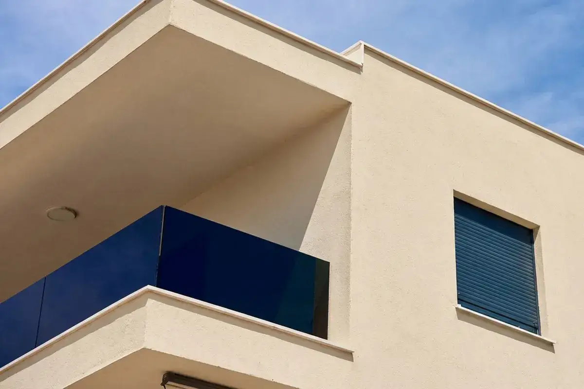 Angolo di una casa moderna con intonaco chiaro, balcone con parapetto blu intenso e finestra con serranda chiusa. Ideale per chi cerca idee per dipingere casa esterno.