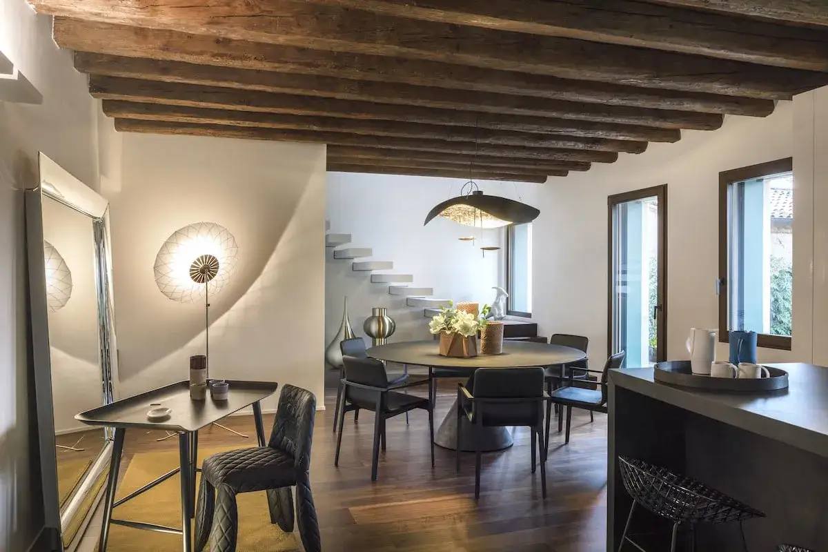 Interno moderno con soffitto con travi in legno a vista, zona pranzo con tavolo rotondo e sedie nere, e un'area salotto con specchio e lampada di design.