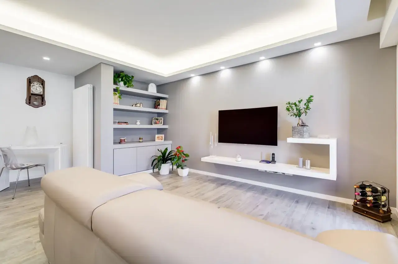 Soggiorno moderno con divano chiaro, parete attrezzata con TV e libreria a giorno, e eleganti ingressi in cartongesso che incorniciano la stanza.