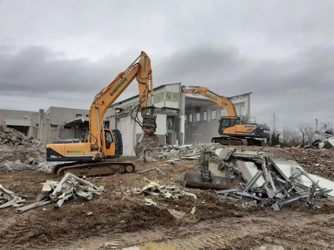 Escavatori gialli in azione, demolendo una struttura e creando cumuli di rifiuti inerti.