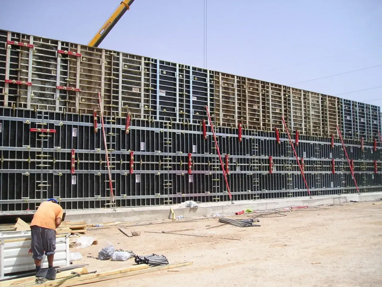 Casse edili in fase di montaggio per un muro di grandi dimensioni, con operai al lavoro e un braccio meccanico sullo sfondo.