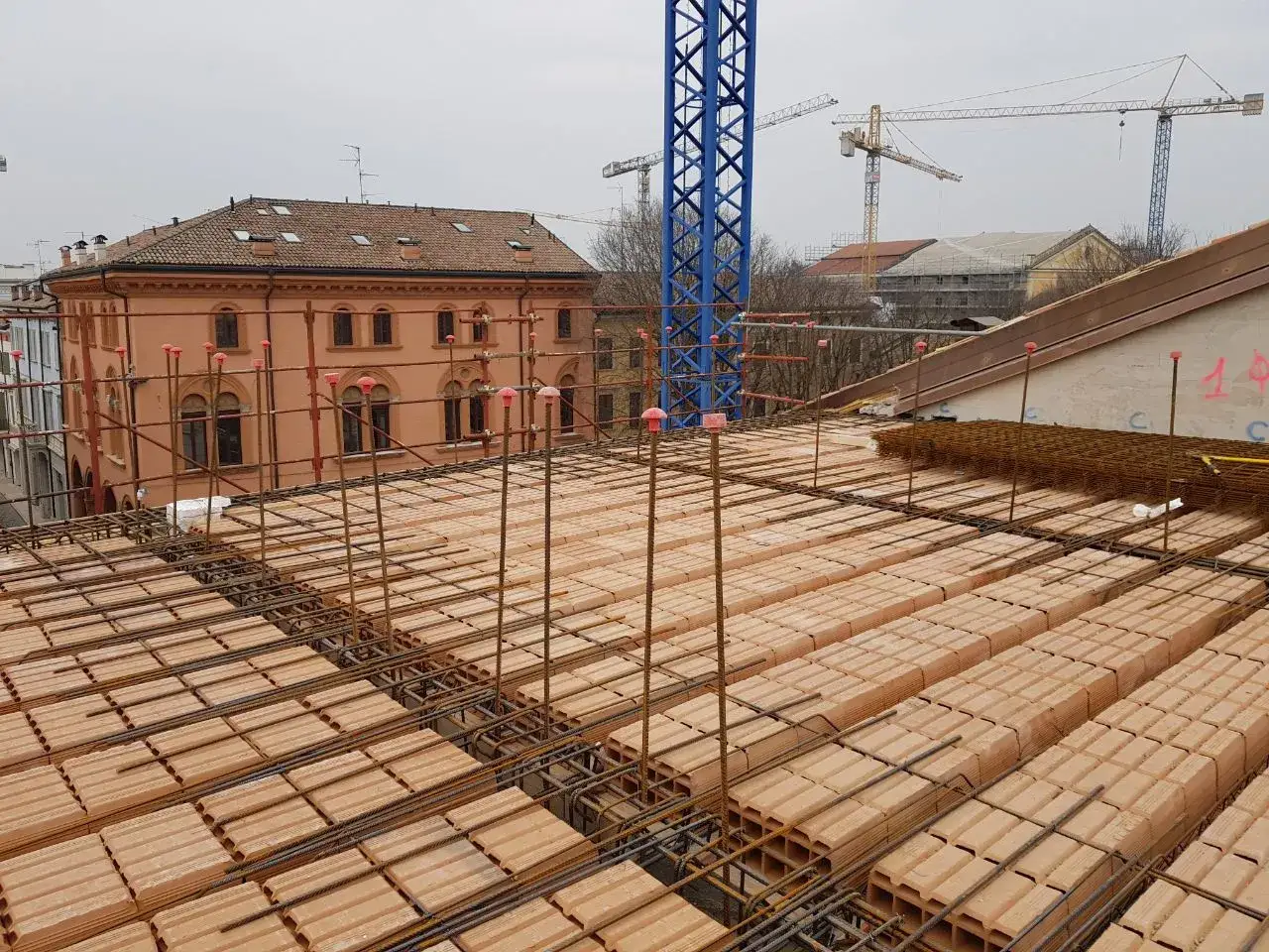 Solaio in laterizio in fase di costruzione, con mattoni rossi e armature metalliche in vista. Gru e edifici sullo sfondo.