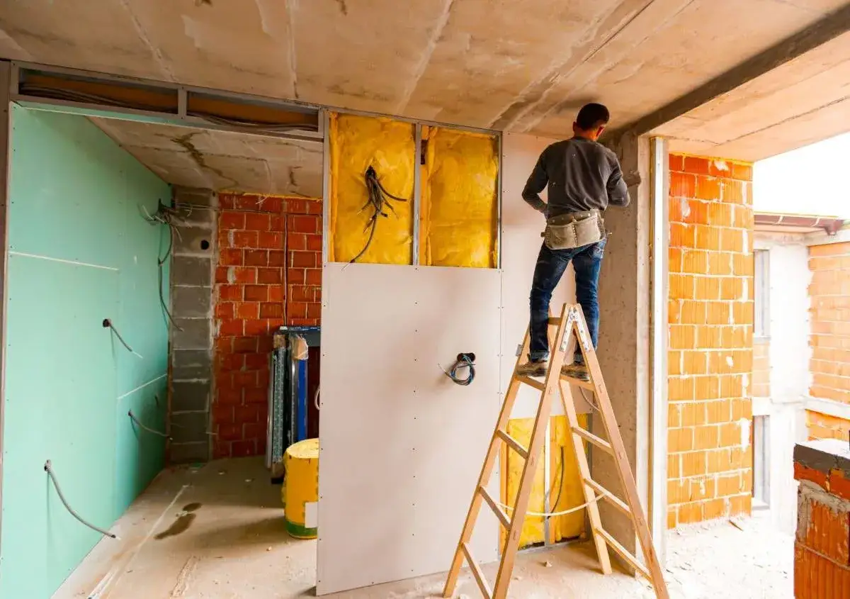 Operaio su scala monta cartongesso, isolamento giallo visibile. Lavori in corso, possibile abbattimento muro interno senza CILA.