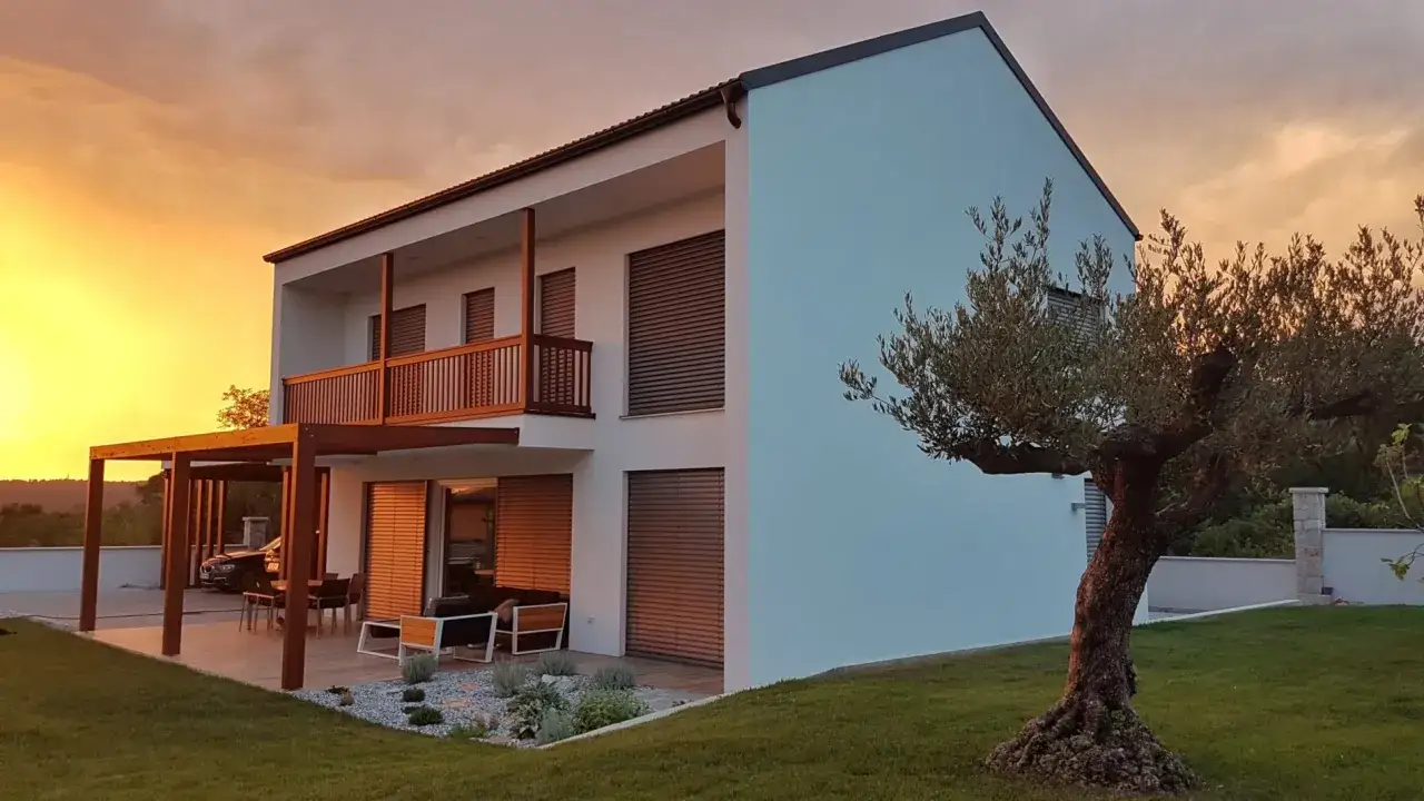 Casa moderna con tettoia in legno e ulivo secolare, esempio di costruzione passiva che abbraccia il tramonto.