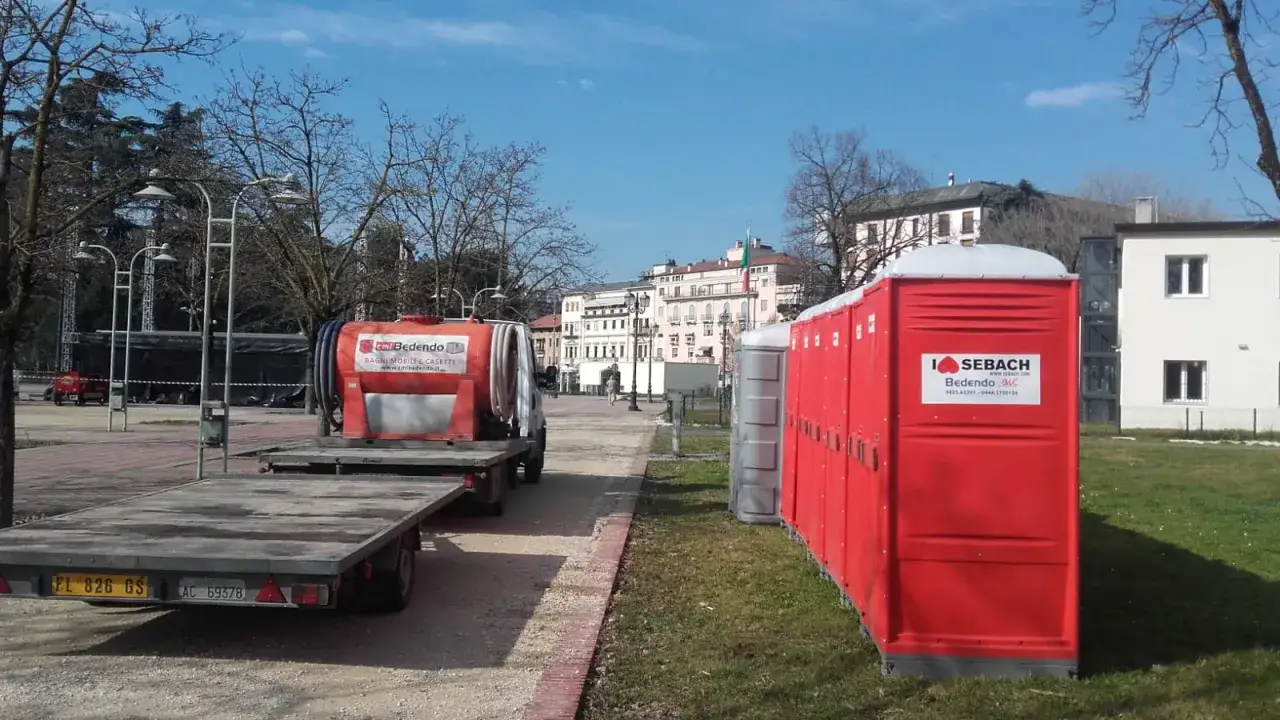 Bagni mobili rossi e grigi allineati, pronti per un evento. Un camion cisterna Bedendo &egrave; parcheggiato nelle vicinanze, suggerendo la gestione dei servizi igienici normativa.