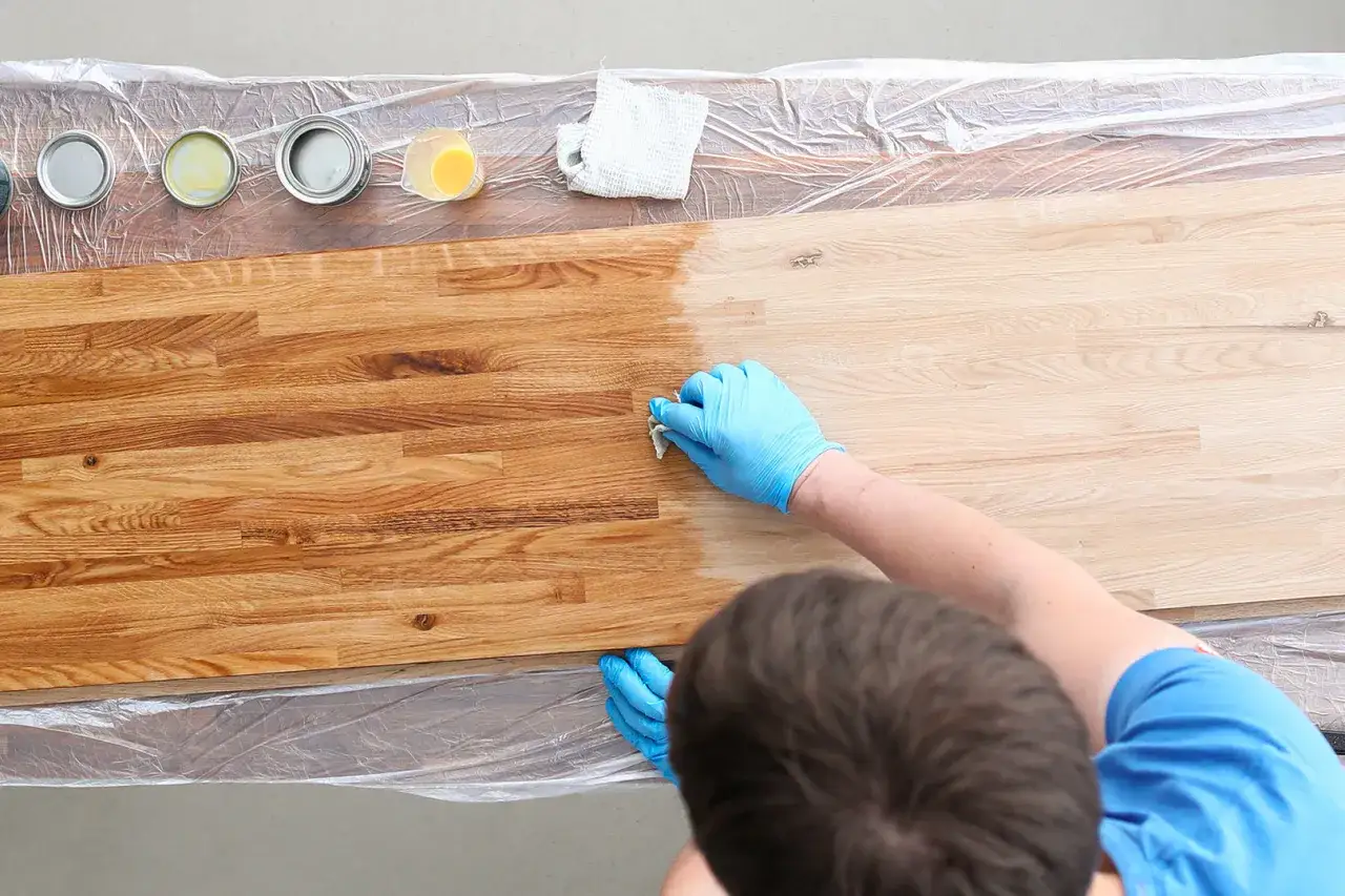 Mani con guanti blu applicano olio per il restauro travi in legno. L'area trattata assume una tonalità più calda e ricca.