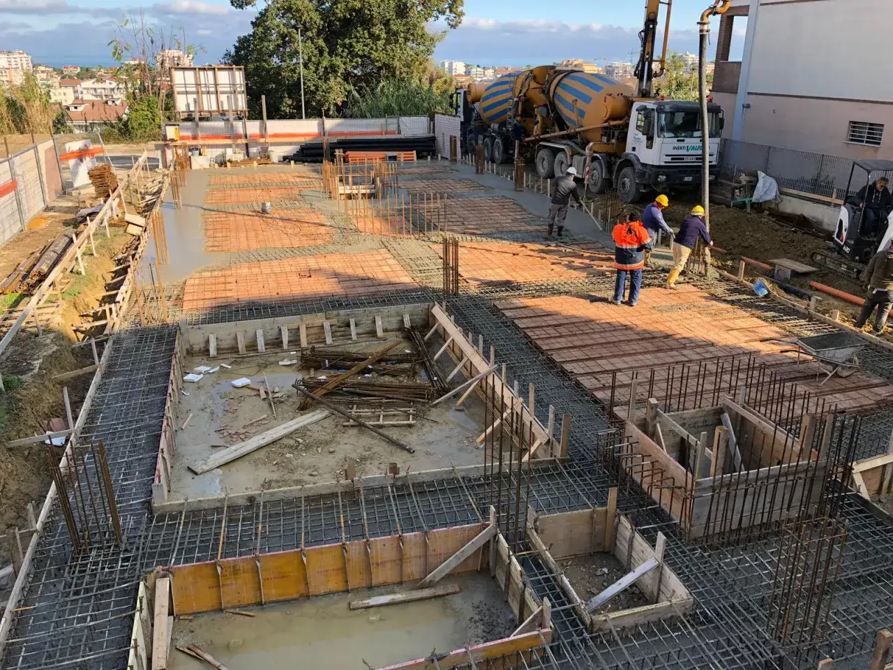 Lavoratori in cantiere versano cemento su una platea di fondazione, con autobetoniera e armature in vista.
