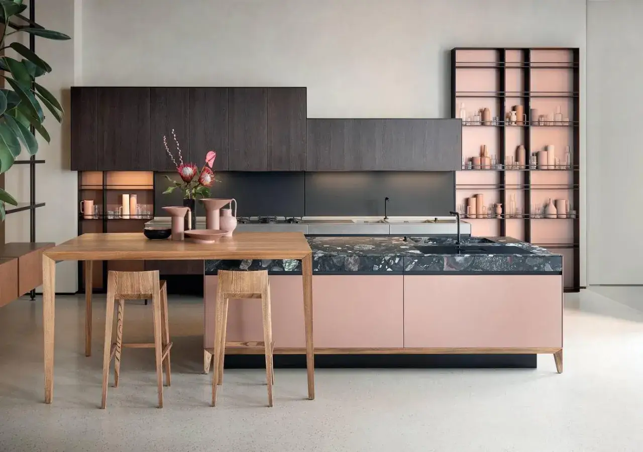 Una cucina moderna con isola centrale color rosa cipria e piano in marmo scuro. L'arredo gioca con il contrasto tra il legno scuro e il colore pastello, creando un ambiente elegante.